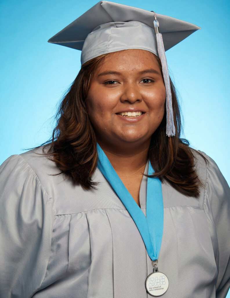 Citlaly Aguayo smiling in a silver graduation cap and gown against a blue background.